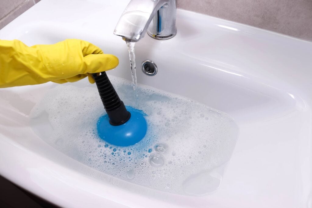 Hand in yellow gloves cleaning bathroom sink with plunger. Overflowing bathroom sink. Clogged drain.