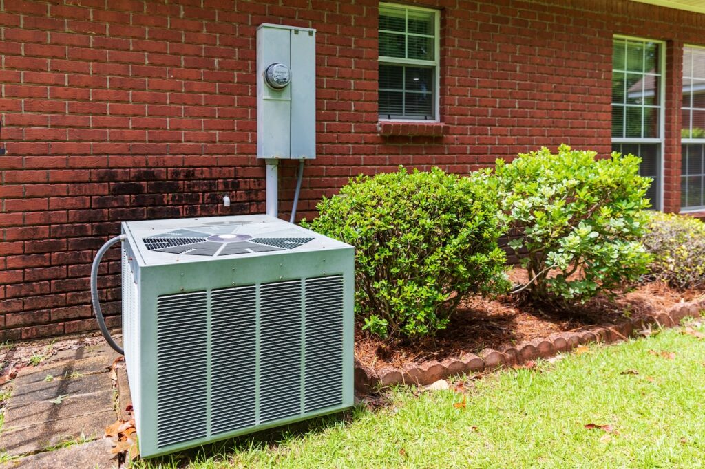 Older style air conditioner unit next to home, brick and bushes with clean yard.